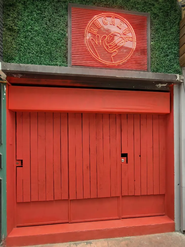 Entrada de Tío Rucahue: puertas rojas de madera y, arriba, el letrero rojo redondo con la mano y la palabra Rucahue sobre un muro de follaje