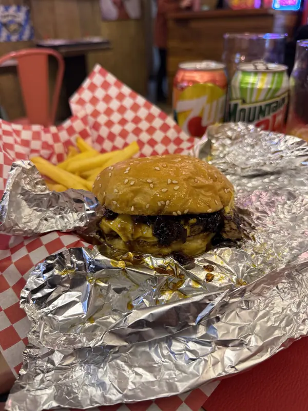 Hamburguesa sobre papel aluminio y papel a cuadros, con latas de 7 Up y Mountain Dew atrás
