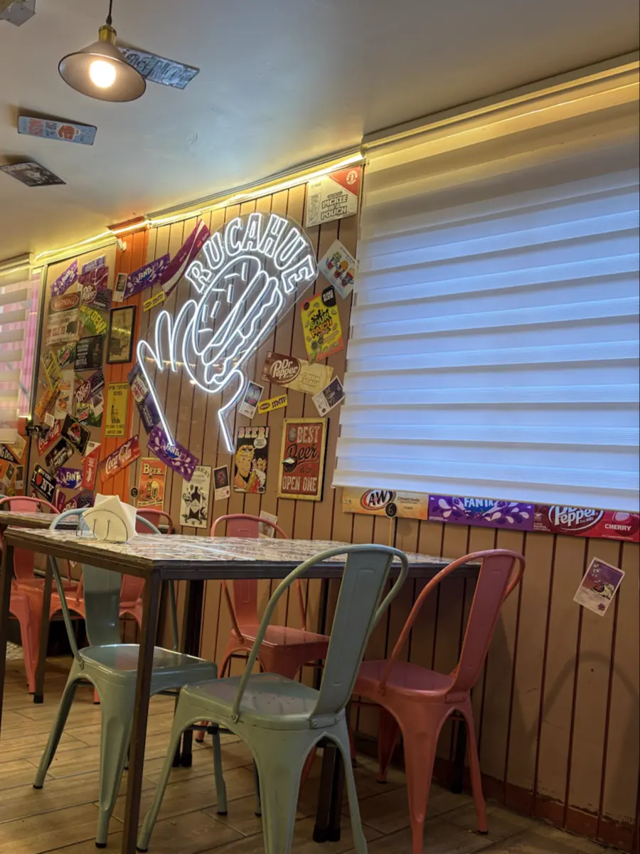 Interior del local: neón blanco con la palabra RUCAHUE y la mano, muros con envoltorios de golosinas, mesas y sillas de metal verde agua y rosadas